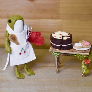 May include: A green felt frog wearing a white apron with a cherry design, holding a whisk and a wooden spoon, stands next to a small wooden table with a chocolate cake and cookies on it.