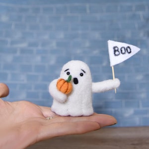 Puede incluir: Un fantasma de fieltro blanco sosteniendo una pequeña calabaza de fieltro naranja y agitando una bandera blanca con la palabra "BOO" escrita en negro.