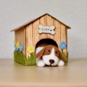 May include: A handcrafted dog house with a felt dog peeking out. The dog house is made of wood-look material, with a bone-shaped sign that says "WOOF". The dog is brown and white, with a red collar. The house is decorated with felt flowers.