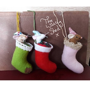 May include: Three felt Christmas stockings with mice peeking out of the top. The stockings are green, red, and pink. The mice are wearing hats and are looking out of the stockings. The text "The Wishing Shed" is written on the background.
