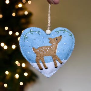 May include: A heart-shaped Christmas decoration with a light blue backdrop and a brown deer with white spots. The ornament features a white snow base and small white embellishments. A beige string is attached for hanging.