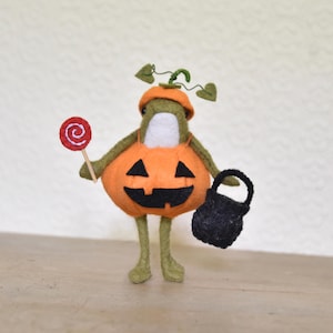 May include: A handmade Halloween decoration featuring a felt frog dressed as a pumpkin. The frog holds a red lollipop and a black trick-or-treat bag. The pumpkin costume is orange with a carved face.
