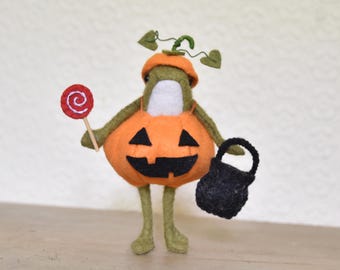 May include: A felted green frog figurine wearing an orange pumpkin costume with a jack-o'-lantern face. The frog holds a red and white swirl lollipop in one hand and carries a small black felt bucket in the other, with a green stem on its head.