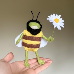 May include: A felt bee toy with a black beard, yellow and brown striped body, green legs, and white wings. The bee is holding a white felt daisy with a yellow centre.