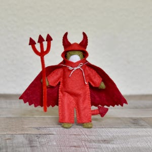 May include: A handmade felt devil doll in a red costume with a matching cape, hood, and tail. The doll holds a red pipe cleaner trident. The doll's costume is secured with a silver string. The doll is standing on a wooden surface.