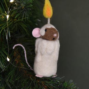 May include: A whimsical Christmas ornament shaped like a mouse candle. The mouse has a brown face, pink ears, and a white body. A yellow and orange flame sits atop the mouse's head. The ornament is set against a backdrop of green pine needles.