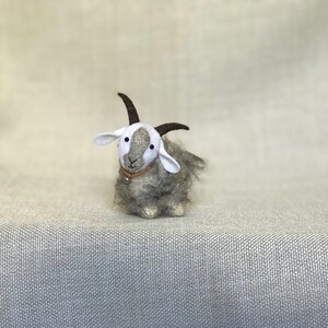 May include: A small gray and white felt goat with brown horns and a brown leather collar with a silver bell. The goat is sitting on a light gray textured surface.