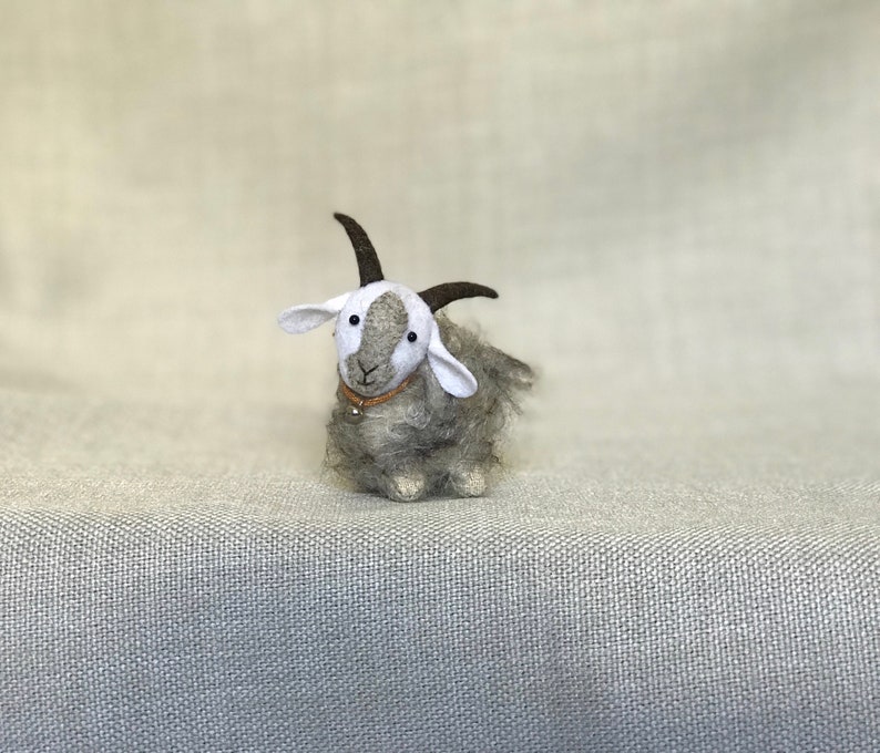May include: A small gray and white felt goat with brown horns and a brown leather collar with a silver bell. The goat is sitting on a light gray textured surface.