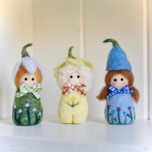 May include: Three felt dolls, each representing a different flower. The first doll is green with white flowers and a white hat. The second doll is yellow with a yellow hat and a flower on its chest. The third doll is blue with a blue hat and purple flowers.