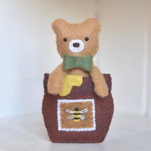 May include: A brown felt bear wearing a green bow tie is peeking out of a brown felt honey pot with a yellow felt honey dipper and a bee embroidered on the front.