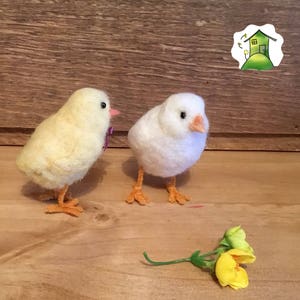 Puede incluir: Dos pequeños pollitos de fieltro, uno amarillo y otro blanco, con patas y picos naranjas. Los pollitos están de pie sobre una superficie de madera con una flor amarilla en primer plano.