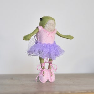 Puede incluir: Una rana de fieltro verde que lleva un tutú de fieltro rosa con una falda de tul morado y zapatos de ballet de fieltro rosa.