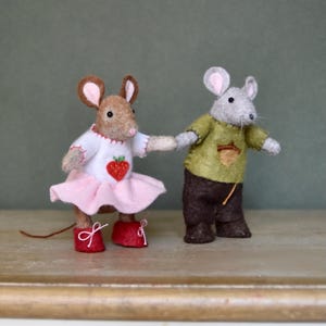 Könnte beinhalten: Zwei handgefertigte Filzmäuse, eine braune und eine graue, halten Händchen. Die braune Maus trägt ein weißes Kleid mit einer Erdbeere und einem rosa Rock, rote Schuhe. Die graue Maus trägt ein grünes Hemd und eine braune Hose.
