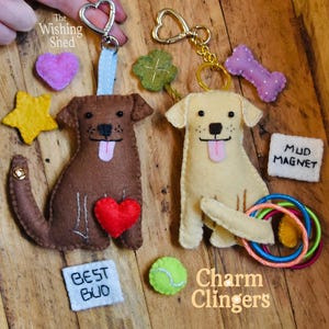 May include: Felt dog keychains with heart-shaped clasps, a brown dog with a red heart, and a yellow dog. Additional charms include a star, bone, clover, and a tennis ball. The text "Best Bud", "Mud Magnet", and "Charm Clingers" are also visible.