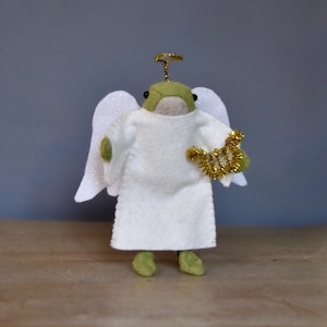 May include: A handmade felt angel figurine with a green head, white robe, and glittery white wings. The angel holds a gold harp and has a gold halo. The figurine is standing on a wooden surface.