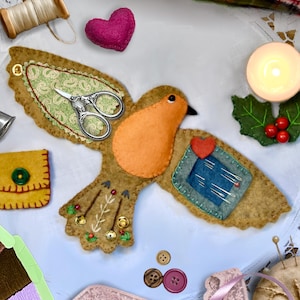 May include: A felt bird-shaped sewing organizer in tan and orange with a green paisley wing, a blue needle storage patch, and decorative embroidery. It features silver scissors, a pink heart, buttons, and sewing supplies arranged around it.