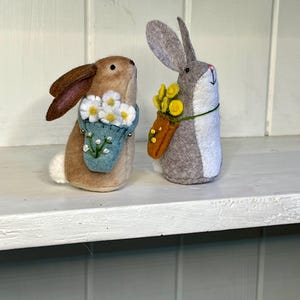 Op de afbeelding: Twee vilten konijnenfiguren. Het bruine konijn draagt een lichtblauw tasje gevuld met witte bloemen. Het grijze konijn heeft een witte borst en draagt een oranje tasje met gele bloemen. Beide hebben geborduurde gezichten.