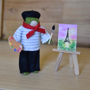 May include: A green felt toy artist wearing a black beret, red scarf, and striped shirt holds a paintbrush and palette. The artist stands in front of a small wooden easel with a painting of the Eiffel Tower.