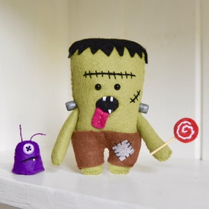 May include: Green felt Frankenstein monster toy with brown felt pants and a purple felt monster toy with one eye. The Frankenstein monster is holding a red felt lollipop.