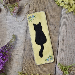 May include: A handmade bookmark featuring a black cat silhouette on a pale yellow felt background. The bookmark has embroidered blue and white flowers and a gold chain for hanging. The edges are stitched with blue thread.