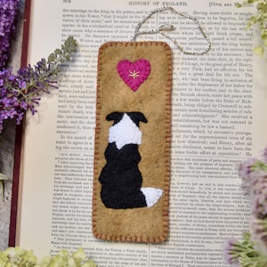 May include: A handmade bookmark featuring a felt Border Collie dog silhouette with a pink heart and gold stitching. The bookmark is rectangular, with a tan background, and is placed on an open book.