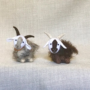 May include: Two small, handmade felt goats. One goat is brown and white, the other is gray and white. Both goats have black eyes and white faces.