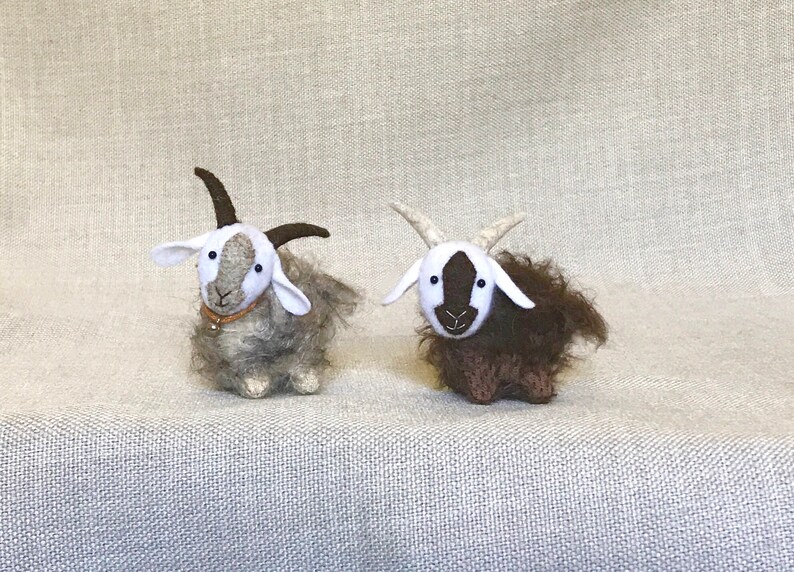 May include: Two small, handmade felt goats. One goat is brown and white, the other is gray and white. Both goats have black eyes and white faces.