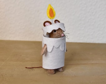 Puede incluir: Un ratón de fieltro hecho a mano disfrazado de vela. El ratón es marrón con orejas rosas y bigotes, vistiendo un disfraz de vela de fieltro blanco con una llama amarilla y naranja. El disfraz de vela tiene un borde festoneado.