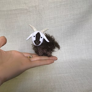 May include: A small, brown and white stuffed goat with a white face and black eyes. The goat is standing on a hand.
