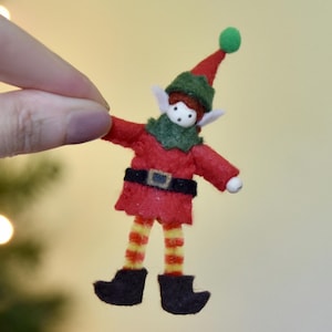 Puede incluir: Adorno de elfo navideño hecho a mano. El elfo viste un atuendo de fieltro rojo con cuello y sombrero verdes, cinturón y zapatos negros, y piernas a rayas amarillas y rojas. El elfo tiene orejas blancas y una cara blanca con puntos negros.