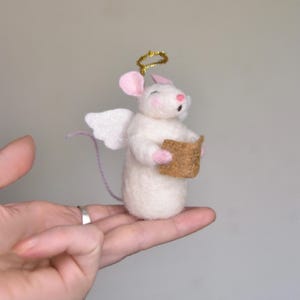 May include: A white felt mouse figurine with pink ears and a pink nose, wearing white glittery wings and a gold halo. The mouse is holding a brown book. A whimsical, handmade Christmas ornament.