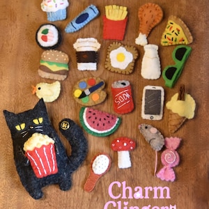 Könnte beinhalten: Eine Sammlung bunter Filzanhänger, darunter ein Cupcake, Sushi, Burger, Pommes, Spiegelei, Pizzastück, eine Limonadendose und eine schwarze Katze mit Popcorn. Der Text "Charm Clingers" ist in Rosa dargestellt.