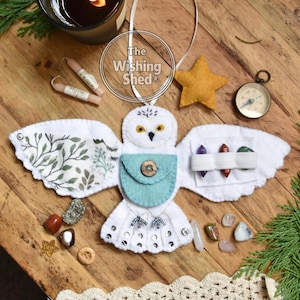 May include: A white felt owl ornament with a teal pouch and button, holding small crystals. The owl has patterned wings and is surrounded by a star, compass, candles, and other crystals. The Wishing Shed logo is visible.