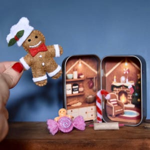 Puede incluir: Un hombre de jengibre de fieltro con gorro de chef y pajarita roja. Una escena navideña en miniatura está dentro de una lata, con un bastón de caramelo y un envoltorio de caramelo rosa con un hombre de jengibre dentro.