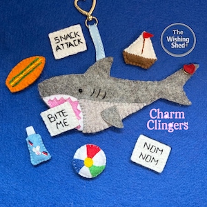 Può includere: Un portachiavi a forma di squalo in feltro grigio con chiusura dorata a cuore, accompagnato da accessori in feltro tra cui una tavola da surf, una barca a vela, un pallone da spiaggia, un tubetto di crema solare e cartelli bianchi con Snack Attack, Bite Me, Nom Nom.