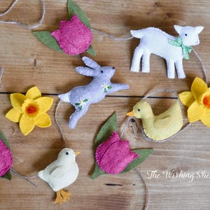 May include: A felt garland featuring a selection of spring-themed decorations, including a yellow daffodil, a pink tulip, a white lamb, a grey bunny, a yellow duck, and a white chick. The garland is strung with twine and has the text "The Wishing Shed" at the bottom.
