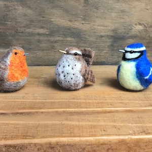May include: Three felt bird figurines, a robin, a wren, and a blue tit, are arranged on a wooden surface. The robin is brown and orange, the wren is brown and white, and the blue tit is blue and yellow.