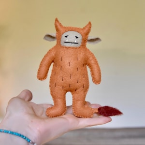 Puede incluir: Muñeca de monstruo de fieltro naranja hecha a mano con capucha con cuernos, orejas pequeñas y cola roja. La muñeca tiene una cara simple con una línea negra para la boca y detalles de costura verticales. La muñeca se sostiene en una mano.