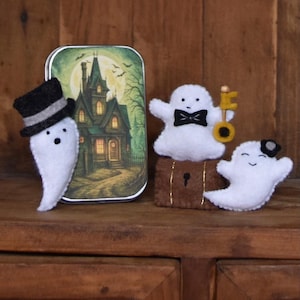 May include: A Halloween-themed display featuring felt ghost figures and a decorative tin. The ghosts are white with black accents, some wearing hats and bow ties. The tin depicts a spooky house scene under a full moon. The items are arranged on a wooden surface.