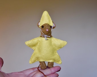 Puede incluir: Un ratón de fieltro hecho a mano que lleva un disfraz amarillo en forma de estrella y un sombrero a juego. El ratón tiene pelaje marrón, orejas pequeñas y una nariz rosa. Una cinta plateada está atada alrededor de su cuello.