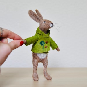 May include: A brown felt rabbit wearing a green felt jacket with a four-leaf clover embroidered on the front. The rabbit has a white patch of fur on its chest.