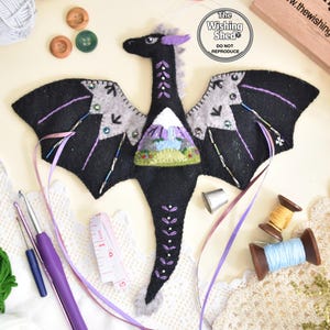 May include: A black felt dragon ornament featuring light grey wing accents, purple embroidery, and a central chest patch depicting a mountain landscape. The dragon is adorned with beads and ribbons, with a logo reading The Wishing Shed.