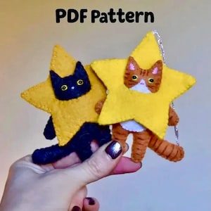 May include: A PDF Pattern featuring two felt ornaments shaped like yellow stars. One star frames a black cat with yellow eyes, and the other frames an orange tabby cat with white markings. Both ornaments have visible stitching along the edges.