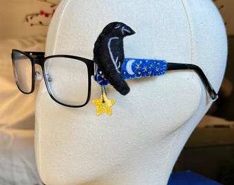 Könnte beinhalten: Schwarz gerahmte Brille mit einem Filz-Raben-Aufsatz am Bügel. Der Bügelüberzug ist blau mit gestickten weißen Mondsicheln und Sternen, akzentuiert durch einen kleinen, baumelnden gelben Glitzerstern-Anhänger.