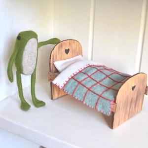 Pode incluir: Uma pequena cama de madeira com um travesseiro branco e um cobertor xadrez azul e vermelho. Um sapo de feltro verde fica ao lado da cama.