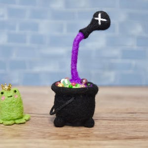May include: A green felt frog wearing a gold crown sits next to a black felt cauldron with a purple felt tube pouring into it. The cauldron is filled with colorful felt balls.