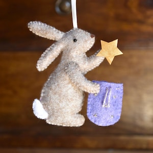 May include: A handmade felt bunny ornament in shades of beige and white, holding a gold star and a purple pouch. The bunny has long ears and a small white tail. A cream-colored ribbon is attached for hanging.