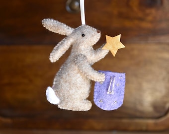 May include: A felt hanging ornament shaped like a beige rabbit with a white tail. The rabbit holds a gold-colored star in one paw and a small purple felt bag with silver embroidered star details in the other. A white ribbon is attached to the top.