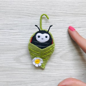 May include: A felt ladybug with a white heart-shaped face and black eyes is nestled inside a green felt leaf with a white flower.
