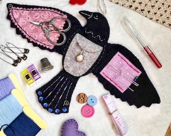 May include: A black felt bird-shaped sewing organizer featuring a pink paisley wing, a gray chest pocket with a pearl charm, and a pink pin cushion on the other wing. It is surrounded by sewing supplies including thread, buttons, pins, scissors, and a tape measure.